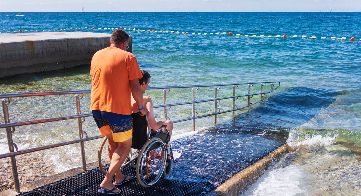 Mann im Rollstuhl, der mit seinem Begleiter über eine Rampe am Strand von Izola ins Meer geht.